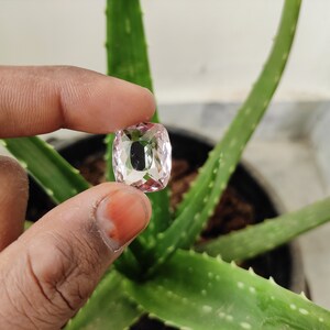 May include: A clear, faceted, pink gemstone with a cushion cut. The gemstone is being held by a hand.