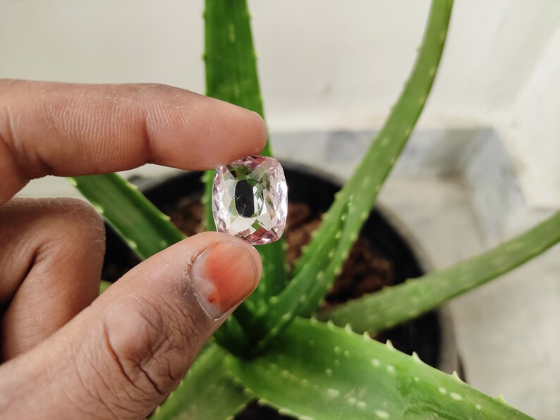 May include: A clear, faceted, pink gemstone with a cushion cut. The gemstone is being held by a hand.