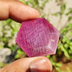 Puede incluir: Una piedra preciosa de rubí facetada de color rosa intenso con forma hexagonal. La piedra preciosa tiene una superficie lisa y pulida y se sostiene en una mano.