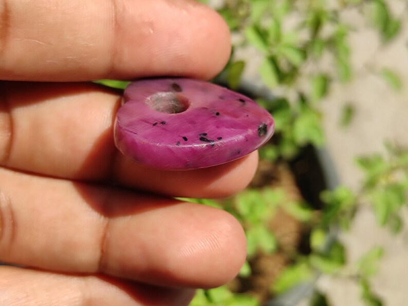 May include: A heart-shaped pink gemstone pendant with black flecks. The pendant has a hole drilled through the top for stringing.