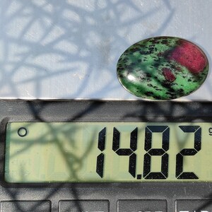 May include: A green and pink oval gemstone with black flecks, weighing 1.482 grams on a digital scale.