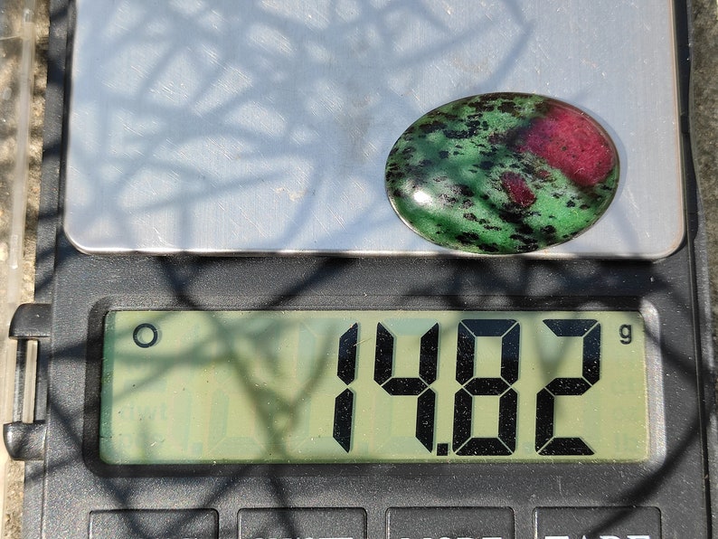 May include: A green and pink oval gemstone with black flecks, weighing 1.482 grams on a digital scale.