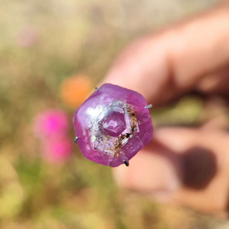 May include: A close-up of a pink gemstone with a hexagonal shape and a silver-colored inclusion in the center. The gemstone is held in place by metal prongs.