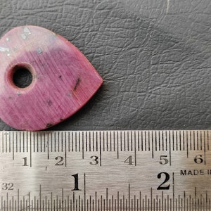 May include: A pink heart-shaped stone with a hole drilled through the centre. The stone is approximately 2.5 cm in length and width. It is shown next to a ruler for size reference.