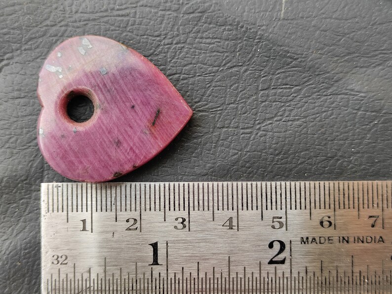 May include: A pink heart-shaped stone with a hole drilled through the centre. The stone is approximately 2.5 cm in length and width. It is shown next to a ruler for size reference.