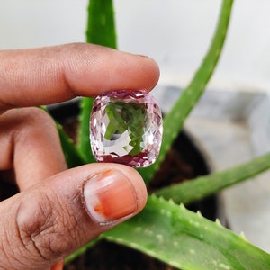 May include: A clear, faceted gemstone with a pink hue. The gemstone is held in a hand, with a green plant in the background.