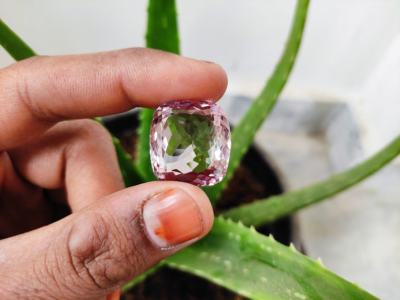 May include: A clear, faceted gemstone with a pink hue. The gemstone is held in a hand, with a green plant in the background.