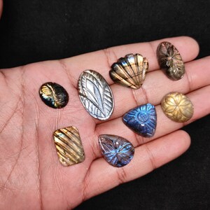 May include: A selection of carved labradorite gemstones in diverse shapes, such as oval, teardrop, and seashell designs. The stones exhibit iridescent hues of blue, gold, and green. The gemstones are presented against a dark backdrop.