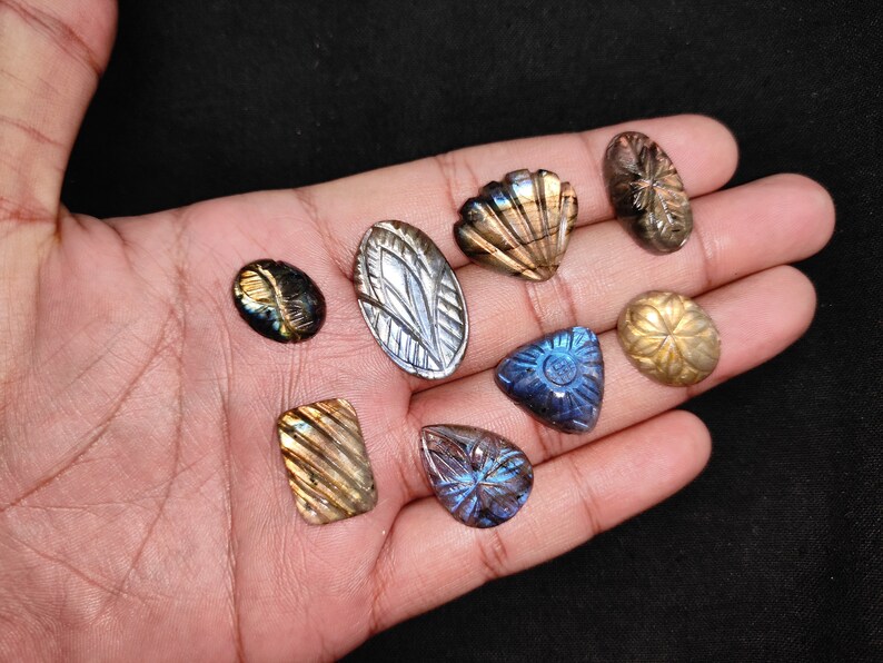 May include: A selection of carved labradorite gemstones in diverse shapes, such as oval, teardrop, and seashell designs. The stones exhibit iridescent hues of blue, gold, and green. The gemstones are presented against a dark backdrop.