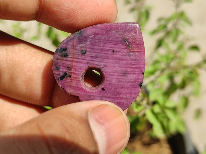 May include: A pink and black heart-shaped gemstone pendant with a hole drilled through the centre. The gemstone has a striated pattern and black inclusions.