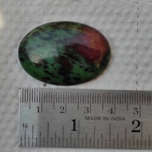 May include: A green and red oval-shaped gemstone with black flecks, measuring approximately 1 inch in length.
