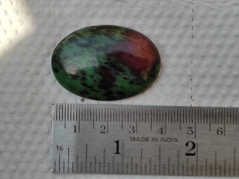 May include: A green and red oval-shaped gemstone with black flecks, measuring approximately 1 inch in length.