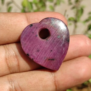 May include: A heart-shaped pendant crafted from pink ruby with a hole drilled through the top. The pendant has a smooth, polished surface and a deep pink colour.