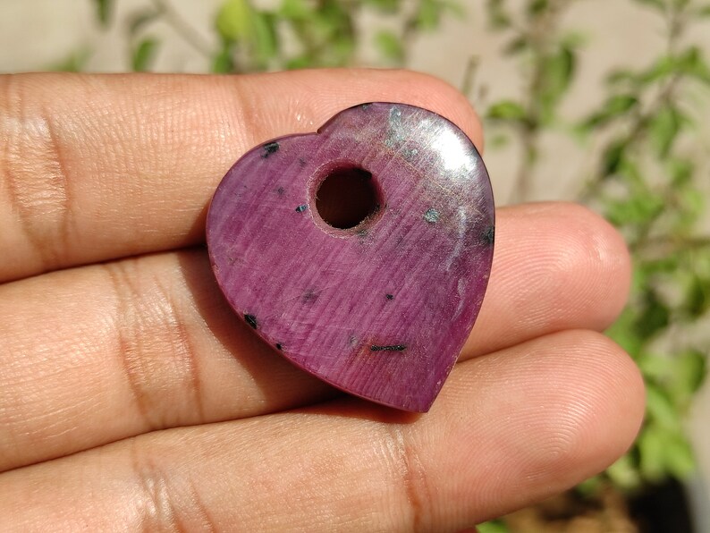May include: A heart-shaped pendant crafted from pink ruby with a hole drilled through the top. The pendant has a smooth, polished surface and a deep pink colour.