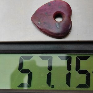 May include: A heart-shaped, pink gemstone with a hole in the centre, weighing 5.715 carats, as shown on a digital scale.
