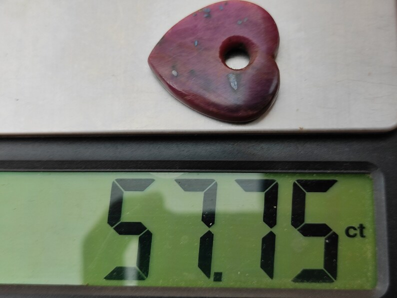 May include: A heart-shaped, pink gemstone with a hole in the centre, weighing 5.715 carats, as shown on a digital scale.