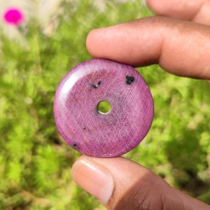 May include: A round, purple gemstone bead with a hole in the center. The bead has a smooth, polished surface and a few black inclusions.