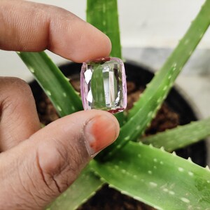 May include: A clear, faceted, pink gemstone with a rectangular shape. The gemstone is being held by a hand against a green aloe vera plant.