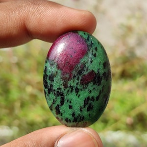May include: Oval-shaped green gemstone with black and pink inclusions. The stone is smooth and polished.