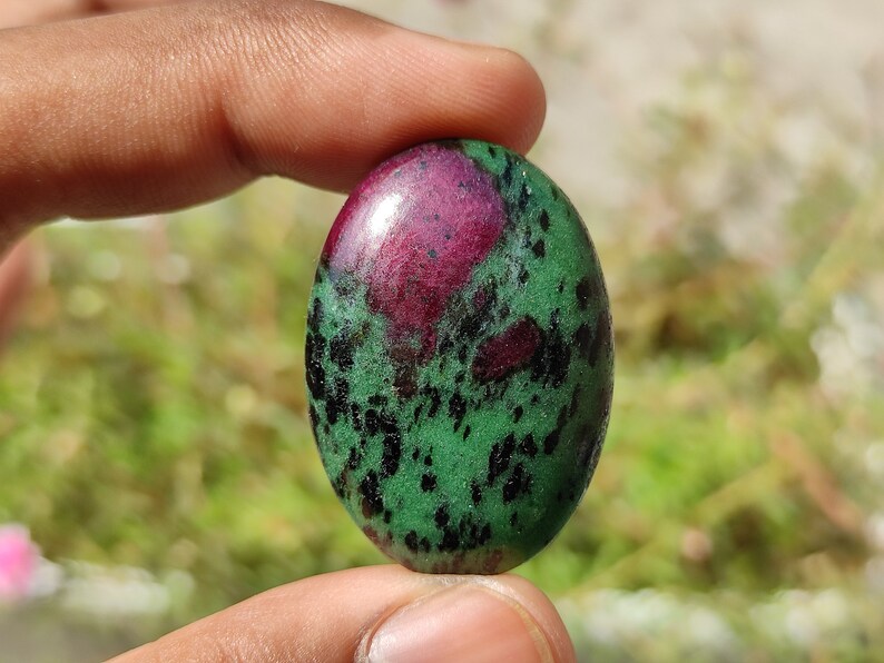 May include: Oval-shaped green gemstone with black and pink inclusions. The stone is smooth and polished.