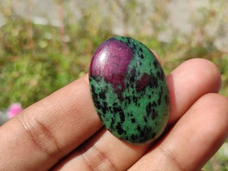 May include: A green and pink oval-shaped gemstone with black speckles. The gemstone is smooth and polished.