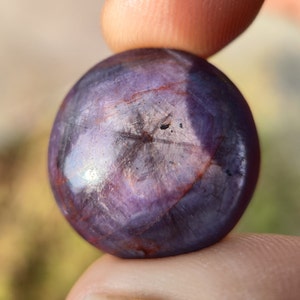 May include: A close-up of a purple and red gemstone with a star-shaped pattern. The gemstone is round and has a smooth, polished surface.