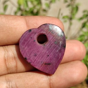 May include: A heart-shaped pendant made of pink ruby with a hole drilled through the centre. The pendant has a smooth, polished surface and a dark pink colour.
