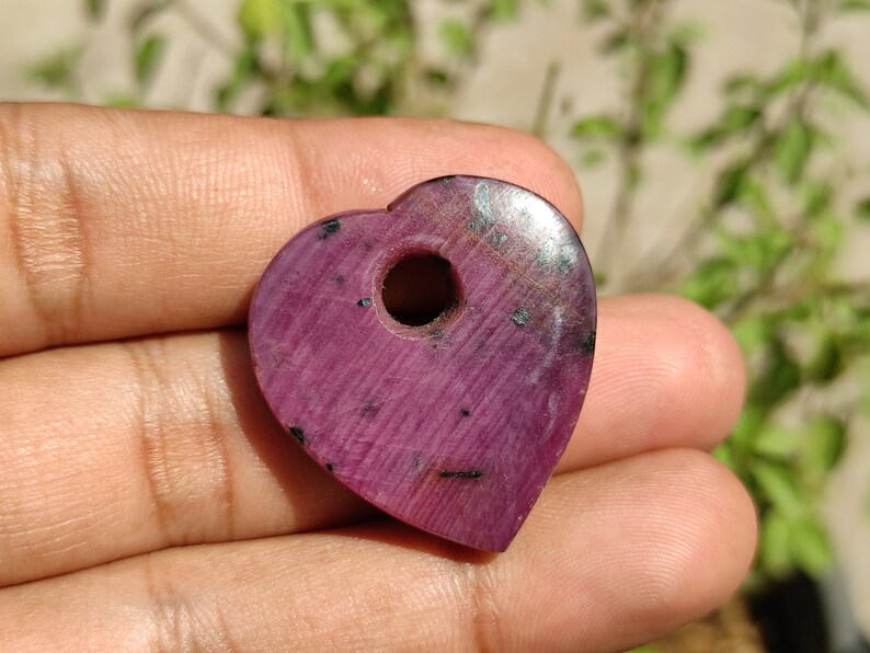 May include: A heart-shaped pendant made of pink ruby with a hole drilled through the centre. The pendant has a smooth, polished surface and a dark pink colour.