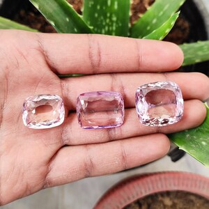 May include: Three pink amethyst gemstones, cut in a cushion shape, are displayed on a hand. The gemstones are different sizes, with the largest one in the center.