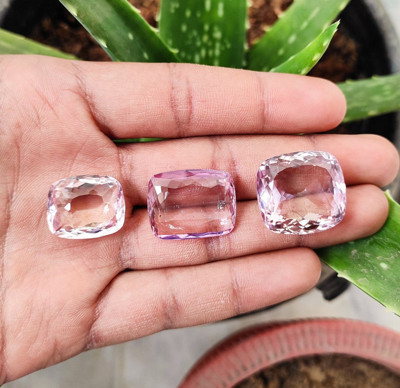 May include: Three pink amethyst gemstones, cut in a cushion shape, are displayed on a hand. The gemstones are different sizes, with the largest one in the center.