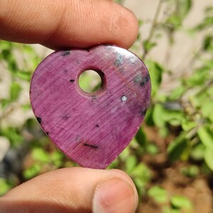 May include: A heart-shaped pendant made of pink ruby with a hole drilled through the top. The pendant has a dark pink colour with black and white inclusions.