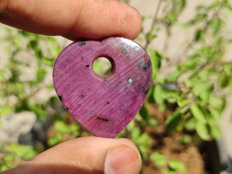 May include: A heart-shaped pendant made of pink ruby with a hole drilled through the top. The pendant has a dark pink colour with black and white inclusions.