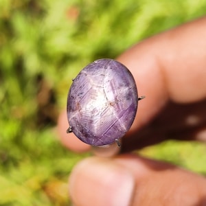 Puede incluir: Una piedra preciosa de zafiro estrellado púrpura con una estrella de seis puntas en el centro. La piedra preciosa está engastada en un engaste de garras de plata.