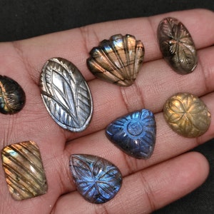 May include: A selection of carved labradorite gemstones in diverse forms, such as oval, teardrop, and rectangular. The stones showcase detailed floral and geometric patterns, with iridescent flashes of blue, gold, and green. The gemstones are presented against a dark backdrop.