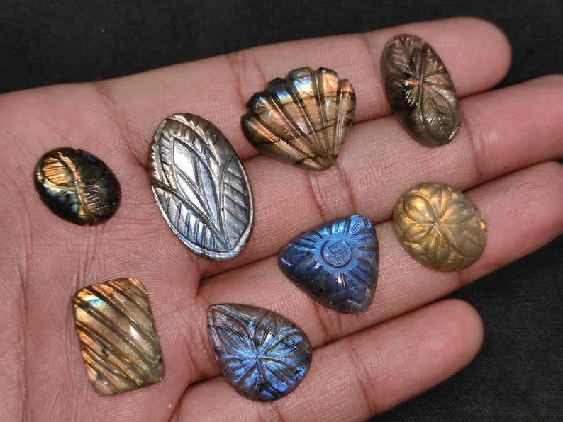 May include: A selection of carved labradorite gemstones in diverse forms, such as oval, teardrop, and rectangular. The stones showcase detailed floral and geometric patterns, with iridescent flashes of blue, gold, and green. The gemstones are presented against a dark backdrop.
