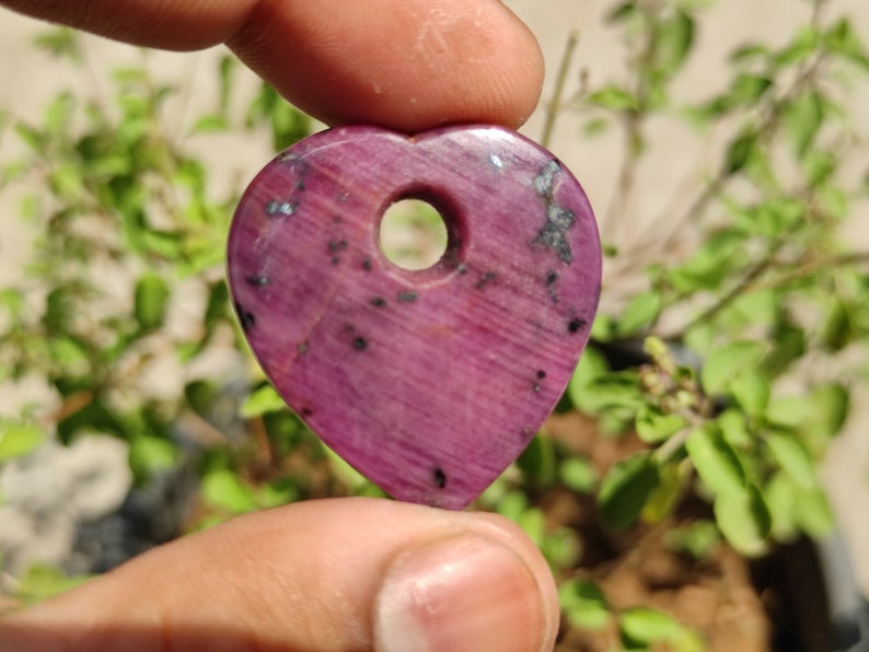May include: A heart-shaped pendant made of pink ruby with black inclusions. The pendant has a hole drilled through the top for stringing.