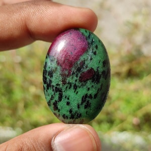 May include: Oval-shaped green gemstone with black and pink inclusions. The stone is polished and has a smooth surface.