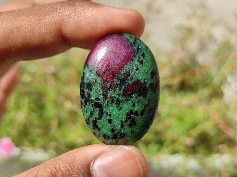 May include: Oval-shaped green gemstone with black and pink inclusions. The stone is polished and has a smooth surface.