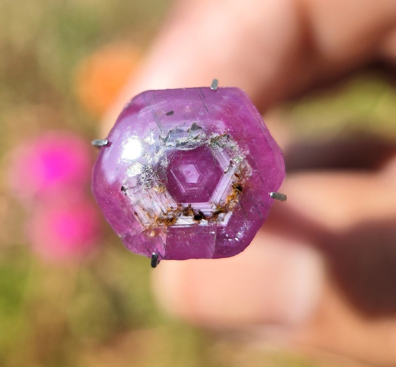 May include: A close-up of a pink gemstone with a hexagonal shape and a clear, faceted center. The gemstone is held in place by metal prongs.