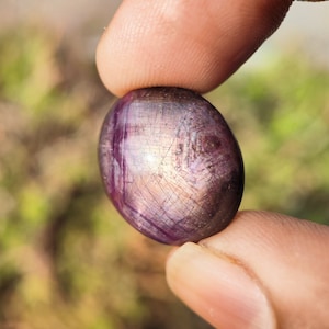 May include: A close-up of a purple gemstone with a smooth, oval shape. The gemstone has a silvery sheen and is held between two fingers.