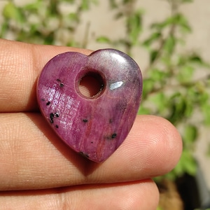 May include: A heart-shaped pendant made of pink ruby with a hole in the centre for a cord or chain. The pendant has a shiny, polished surface and a few black inclusions.