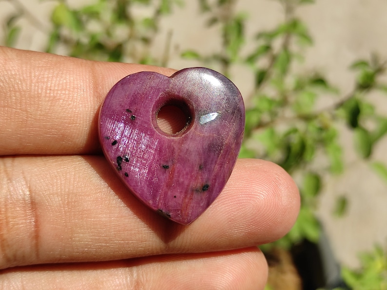 May include: A heart-shaped pendant made of pink ruby with a hole in the centre for a cord or chain. The pendant has a shiny, polished surface and a few black inclusions.