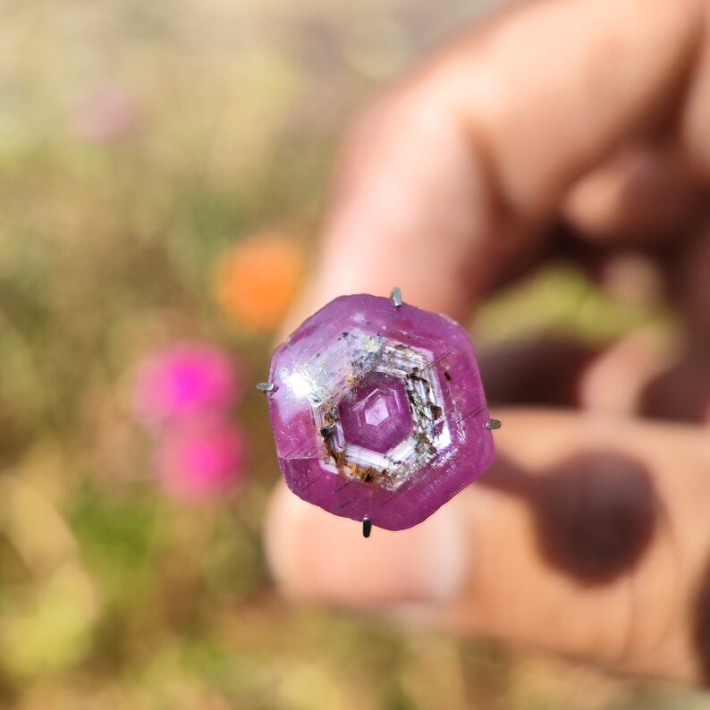 May include: A close-up of a pink gemstone with a hexagonal shape and a silver-colored center. The gemstone is held in place by prongs.