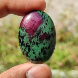 May include: Oval-shaped green gemstone with black and pink inclusions. The gemstone is polished and has a smooth surface.