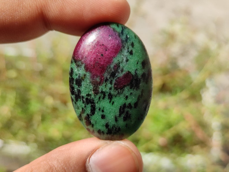 May include: Oval-shaped green gemstone with black and pink inclusions. The gemstone is polished and has a smooth surface.