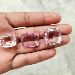 May include: Three faceted gemstones, one clear and two pale pink, are displayed on a hand. The gemstones are cut in a cushion shape.