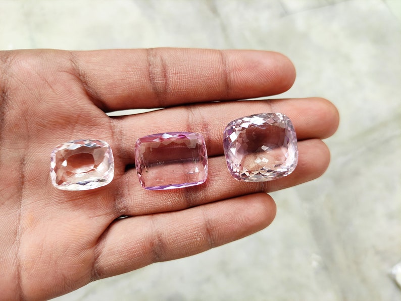 May include: Three faceted gemstones, one clear and two pale pink, are displayed on a hand. The gemstones are cut in a cushion shape.