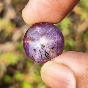 May include: A round, purple gemstone with a star-shaped pattern and black inclusions. The gemstone is held in a hand.