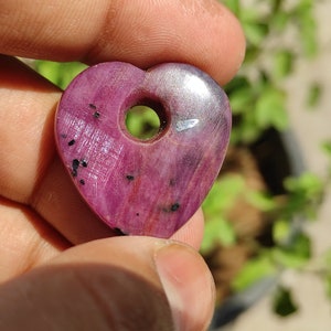 May include: A heart-shaped pink ruby pendant with a hole drilled through the top. The pendant has a silver-coloured backing and black inclusions.