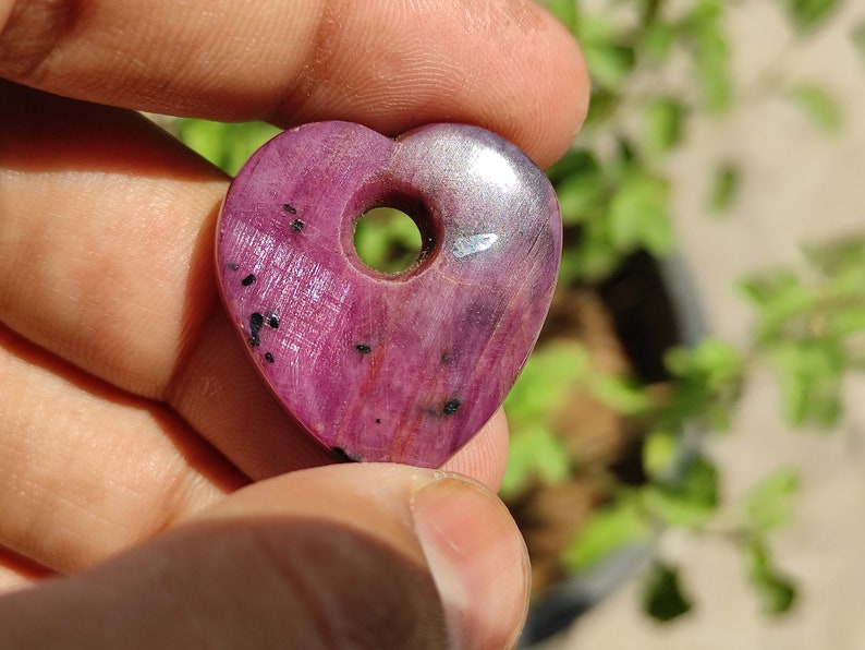 May include: A heart-shaped pink ruby pendant with a hole drilled through the top. The pendant has a silver-coloured backing and black inclusions.
